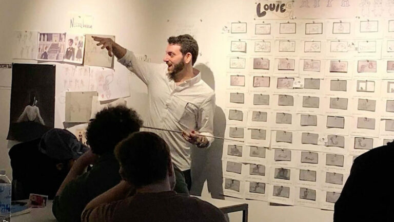 Person presenting in front of an audience, pointing at a wall covered with sketches and storyboard images.
