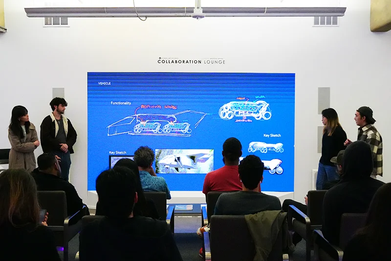 Presenters show vehicle sketches on a large screen labeled "Collaboration Lounge" to an audience in a classroom. Key sketches and functionality appear in bright colors against a blue background.