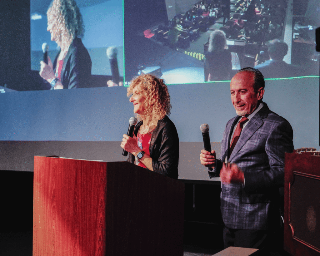 Two people hold microphones, speaking at a podium in a conference setting. A large projection screen behind them displays their images.