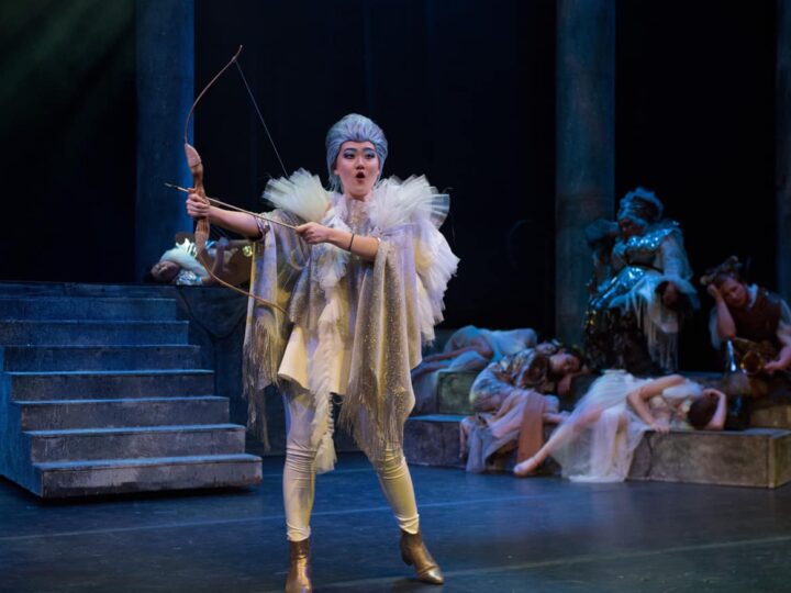 A performer in theatrical costume holds a bow and arrow, appearing focused. The stage has steps on the left and other performers rest in the background, suggesting a drama scene.