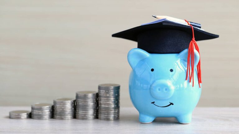 A blue piggy bank wears a graduation cap, symbolizing education savings. It's placed next to stacked coins on a light wooden surface, emphasizing financial growth and academic aspirations.